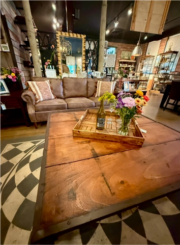 Boutique interior seating area at the Fortin Ironworks Event Lounge featuring a leather sofa, reclaimed wood coffee table, and fresh flowers surrounded by The Market's curated inventory